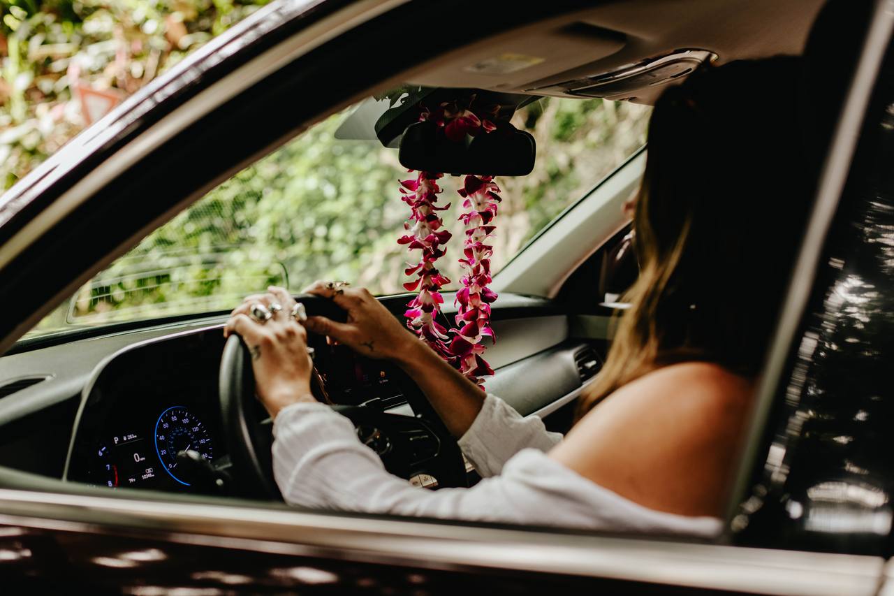 Driver behind the wheel of a Makoa rental car with a fresh orchid lei on the rear-view mirror, Maui